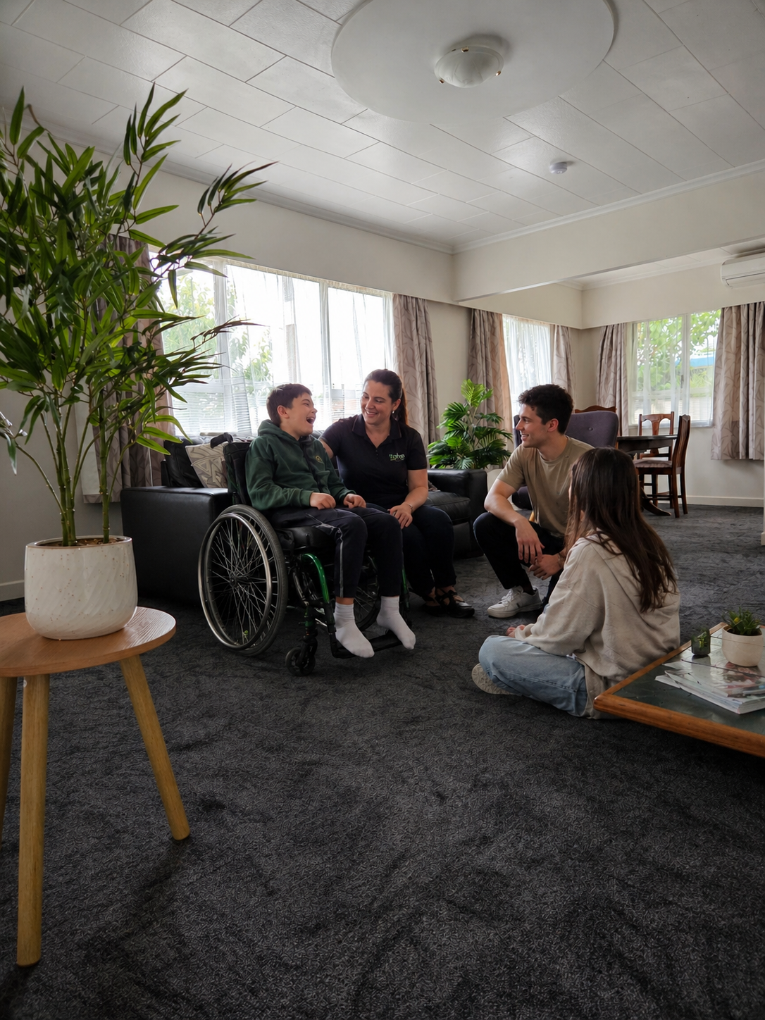 Family gathered in a bright, accessible living room, with a young wheelchair user, caregiver, and siblings relaxing together.