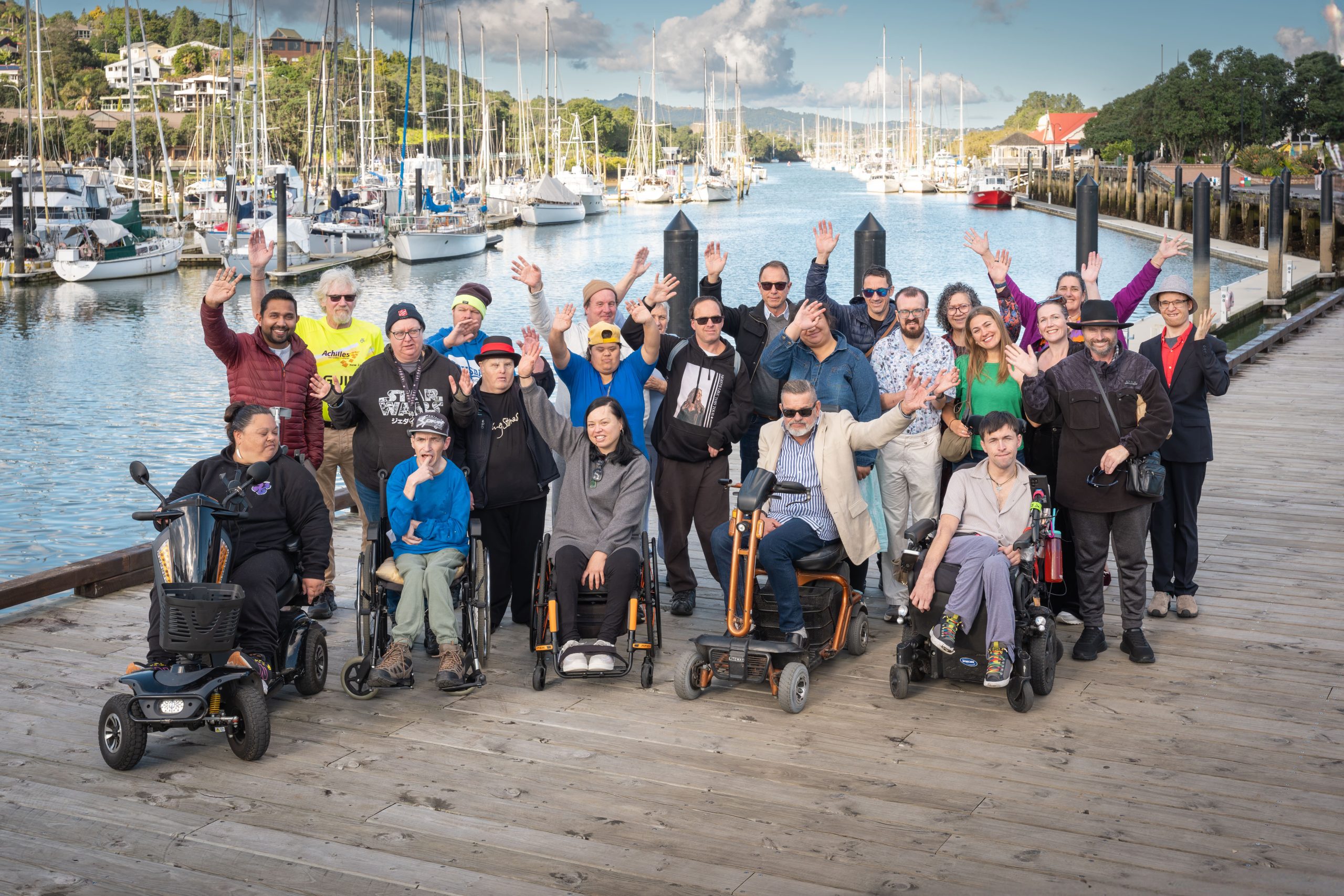 A diverse group of people, including wheelchair and mobility scooter users, gathered on the waterfront at Whangārei Town Basin.